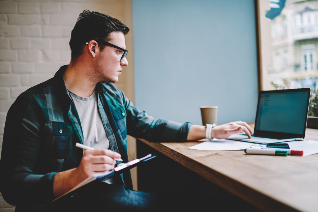 man writing in cafe while laptop in front